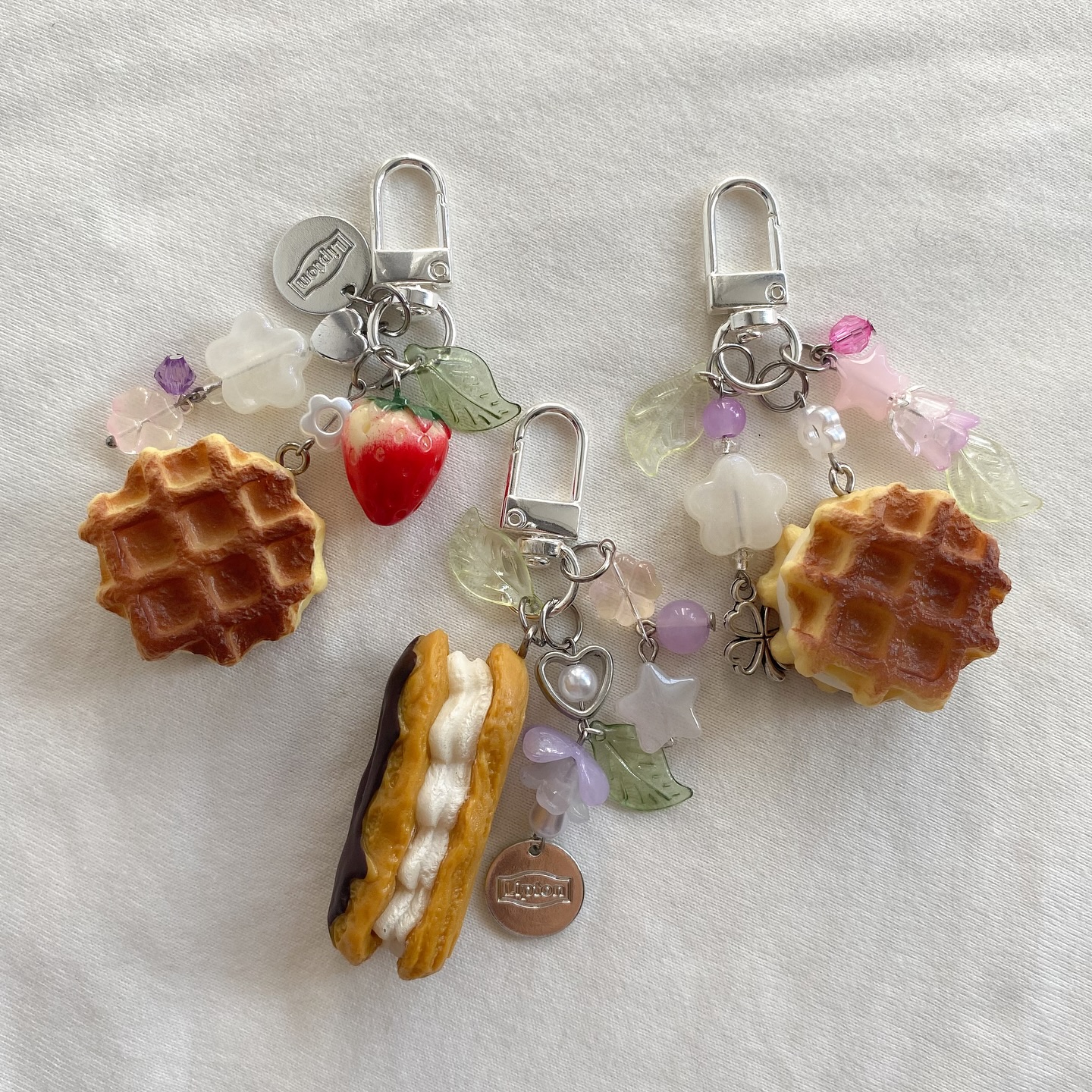 the image shows three cute keychains with a food theme, laid out on a white textured surface. each keychain has a silver clip at the top. one keychain has a waffle charm, a strawberry charm, beads and a small tag with text. another keychain has a waffle charm, floral beads, and transparent green leafshaped beads. the last keychain features an eclair charm, heartshaped charm, small tag with text, and various beads and leaf charms.