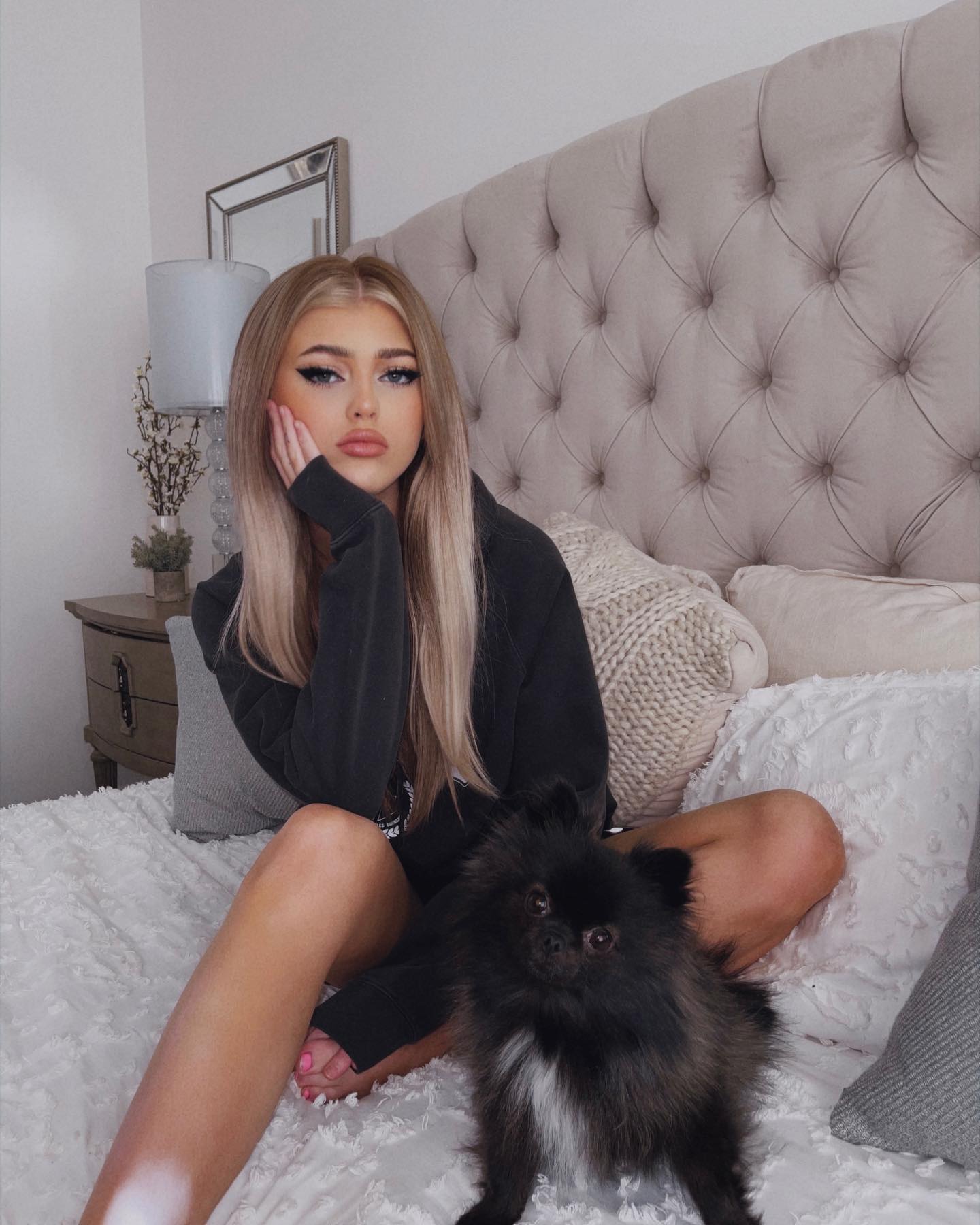 a young woman with blonde hair is sitting on a bed with a black pomeranian dog. she is wearing a black hoodie and has her hand on her face. the bed has a large, tufted headboard and several pillows. there is a lamp and a nightstand next to the bed. the background is blurred.