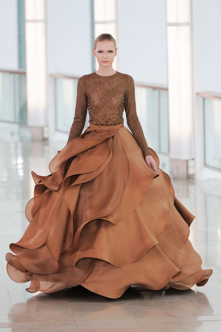 a woman in a brown gown with ruffles walks down a runway. the gown is a rich brown color and has a fitted bodice and a flowing skirt. she has a simple hairstyle and a natural makeup look. the runway is a white tiled floor and the background is a white wall with windows. the woman is looking at the camera and has a confident expression on her face.