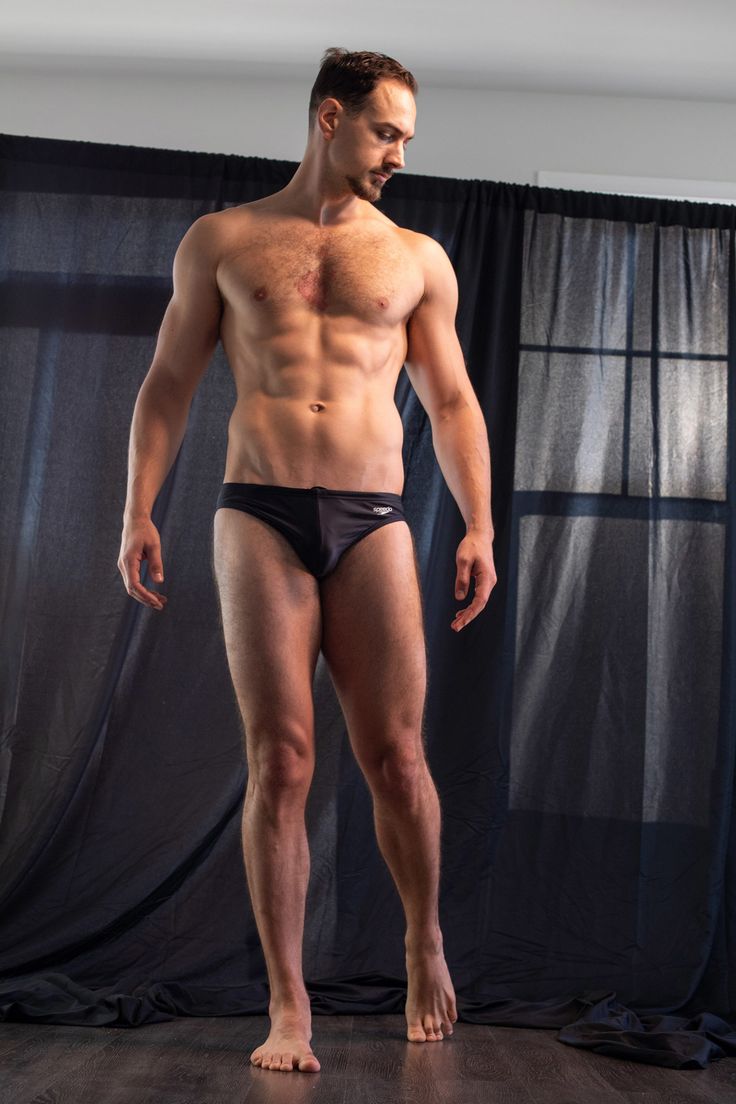 a muscular man is standing in front of black curtains, wearing a black swimsuit. he is looking down and has his arms by his side. the man is barefoot and standing on a dark wooden floor. he is muscular and athletic, with defined abs and pecs.  the image is a portrait shot.