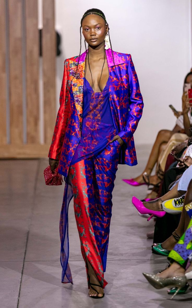 a female model walks the runway wearing a vibrant and colorful pantsuit. the outfit features a mix of blue and red silk fabrics with intricate patterns and textures. she is accessorized with statement earrings and a necklace, and her hair is styled in long braids. the background shows other attendees of the fashion show. the overall impression is one of high fashion, showcasing bold africaninspired designs.