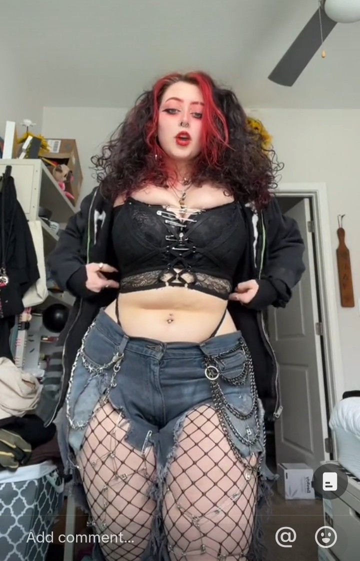 a full body shot of a woman with dark, curly hair and red highlights is standing in a bedroom. she is wearing a black lace top, ripped denim shorts with chains, and fishnets. she also has on a black hoodie that is slightly open, and she has a belly piercing. her hair is pulled back into pigtails.