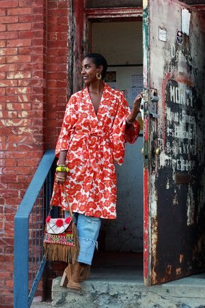 a stylish woman stands in front of a doorway. she is wearing a red and white floral kimono, distressed denim jeans, and brown ankle boots. she carries a fringed handbag and is accessorized with earrings. the background consists of a weathered brick wall and a rusty metal door. the overall aesthetic is bohemian and eclectic, reflecting urban street style.