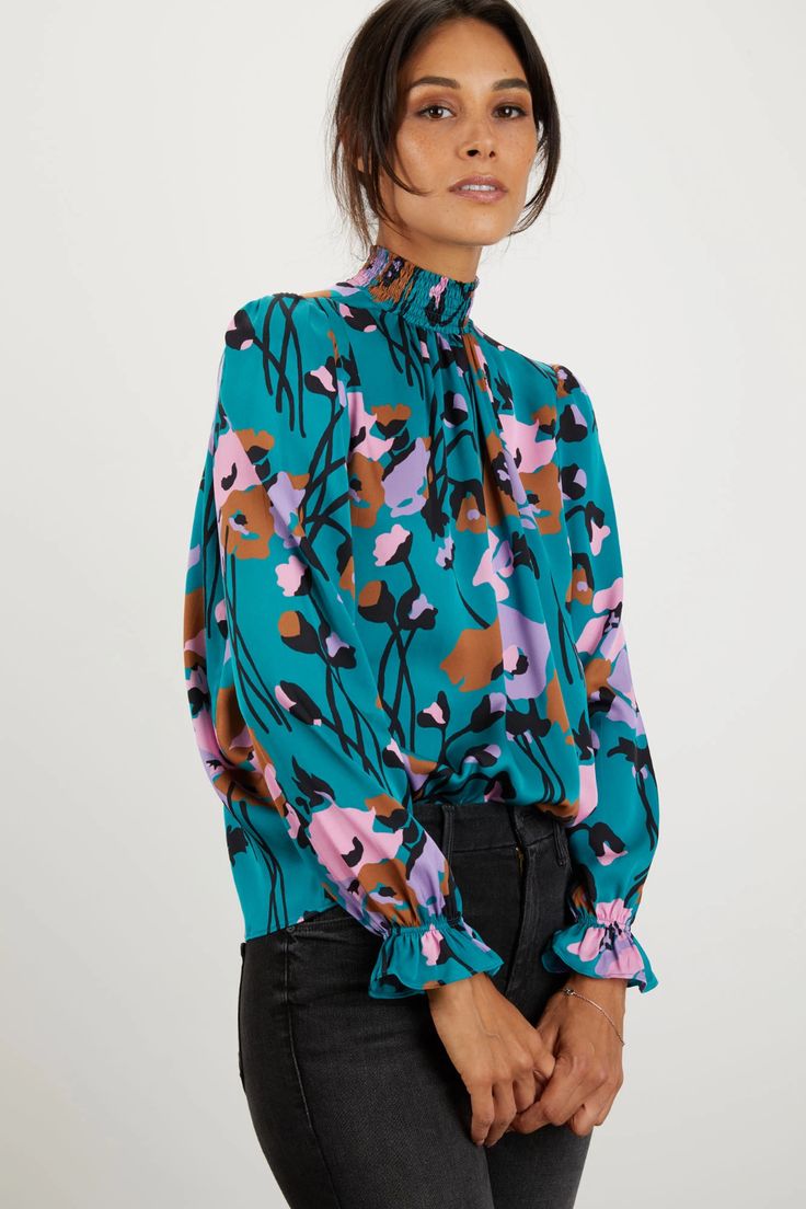 a woman is modeling a teal floral blouse with a high neck and ruffled cuffs. the blouse features an abstract floral print in shades of pink, brown, and purple. the model is wearing dark grey jeans and has her dark brown hair pulled back. the background is a simple white.