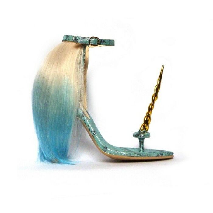 a single unicornthemed high heel shoe is displayed against a white background. the shoe is light blue with a textured pattern reminiscent of scales, featuring a golden, spiraled unicorn horn in place of a traditional heel. a strand of hair, blonde at the top and transitioning to blue at the bottom, drapes down from the shoe's ankle strap, mimicking a unicorn's mane or tail. the shoe has a delicate ankle strap with a buckle.