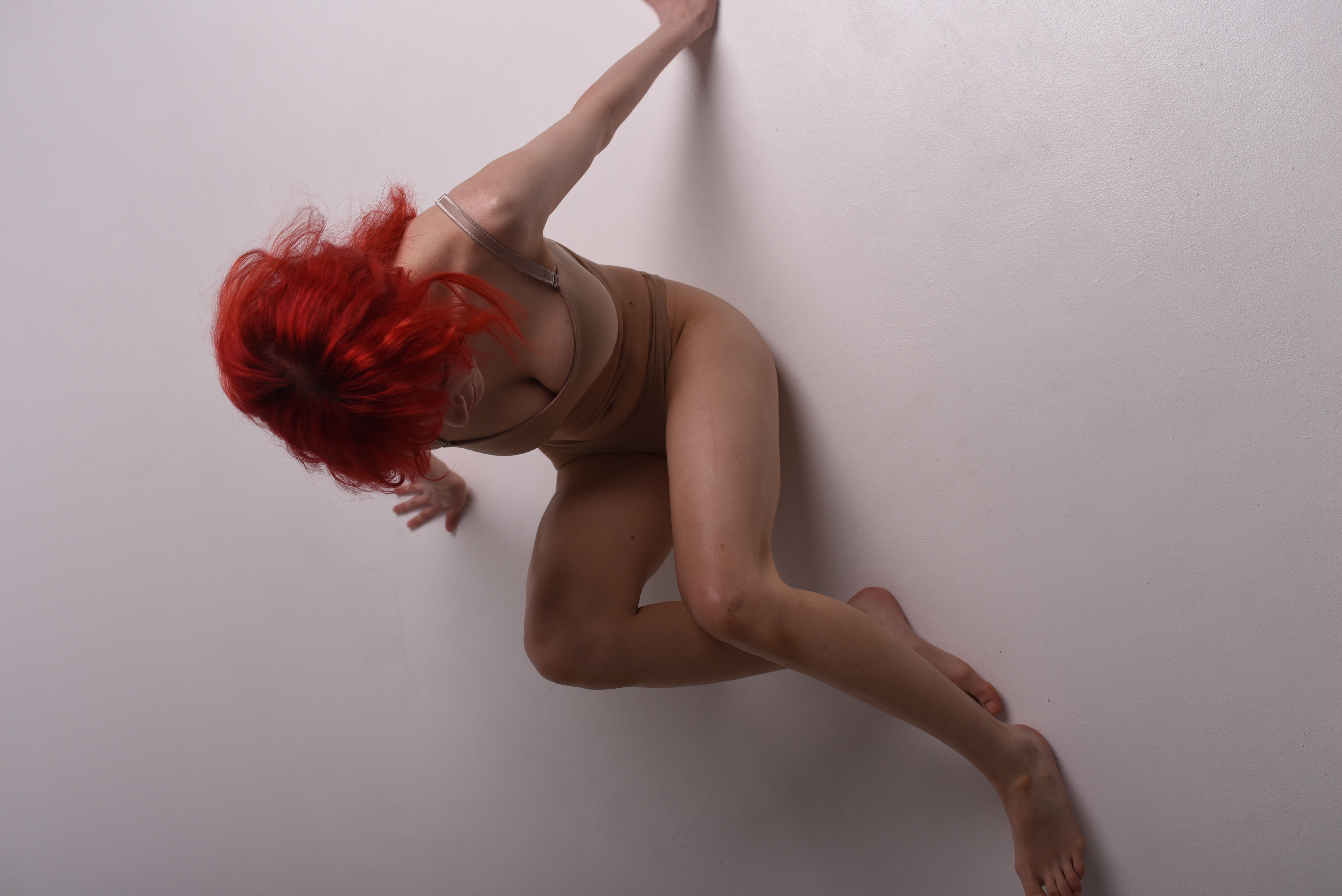 a woman with red hair is laying on a white studio floor. she is wearing a nudecolored bodysuit and her limbs are positioned in a dancelike pose.