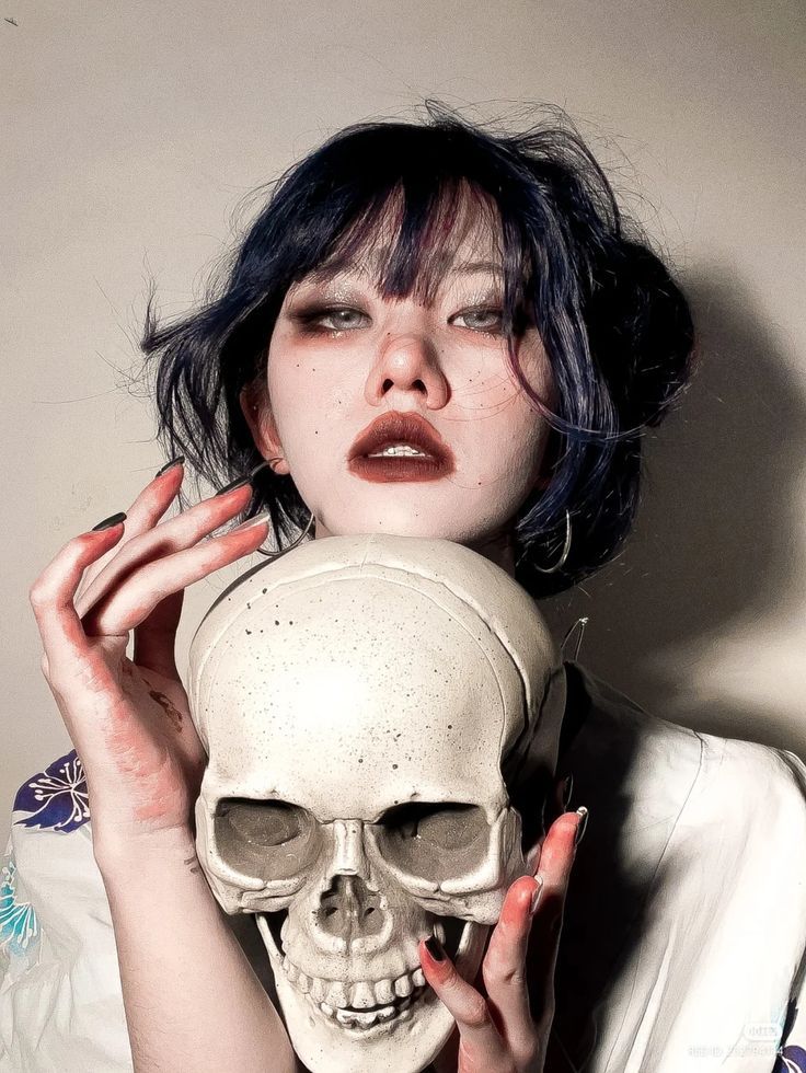 a young woman with dark hair and dark eye makeup holds a white skull in front of her face. she is dressed in a white shirt with a floral pattern on the sleeve. her face is partially obscured by the skull.