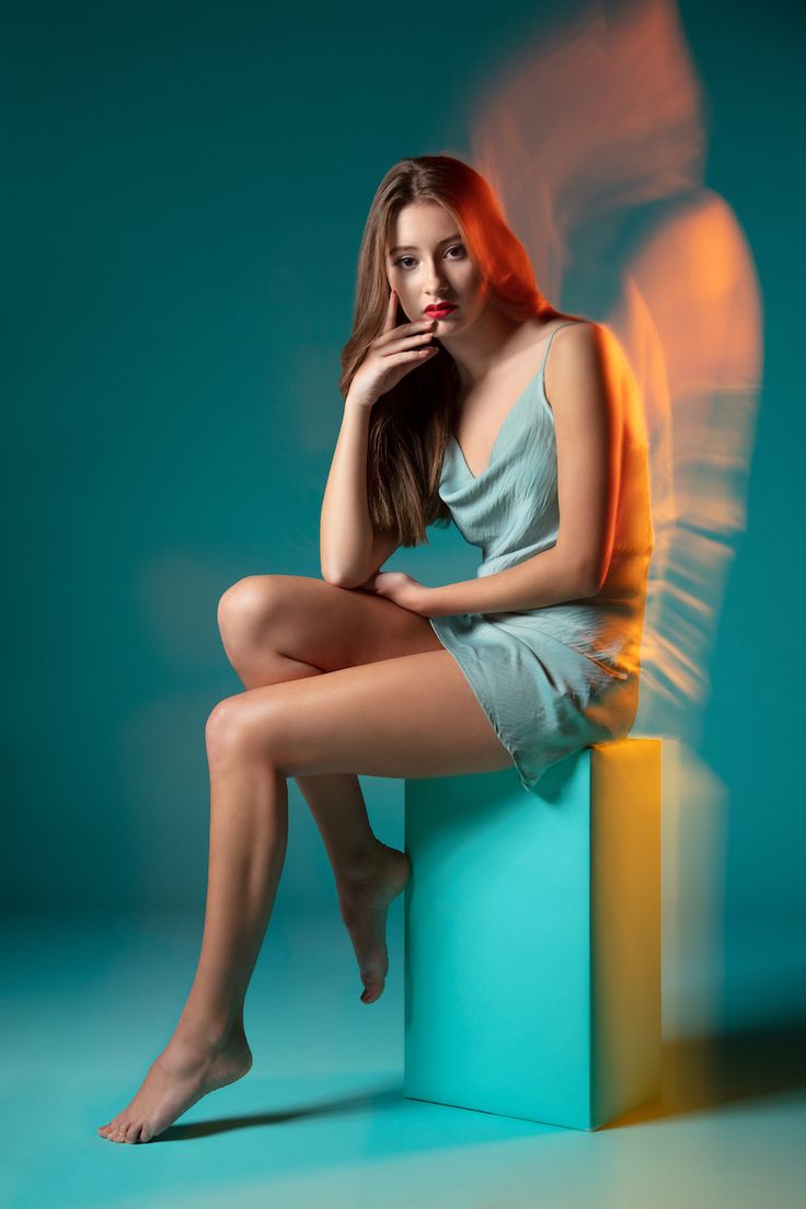 a young woman with long brown hair is sitting on a blue box. she is wearing a light blue dress.  she is looking at the camera. there is a orange light in the background. the woman is  wearing red lipstick. she is a beauty model. the background is blue and the light is casting a shadow on the wall. the woman is sitting on the edge of the box with her legs crossed.  the woman is posing for a photo.