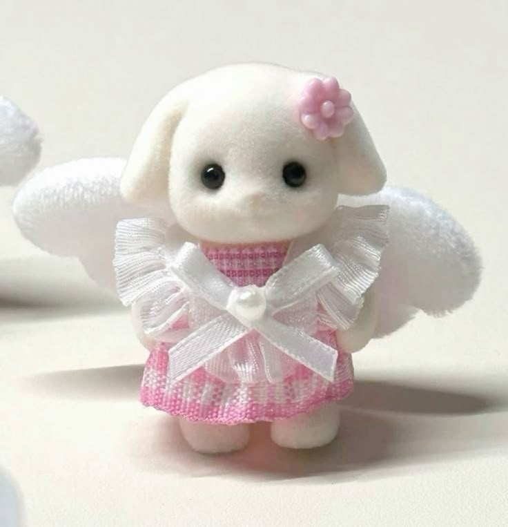 a calico critters toy of a white dog figurine dressed as an angel is pictured. the dog has soft white fur and large, black eyes. it wears a pink plaid dress with a white, frilly overlay and a bow tied at the front with a pearl in the center. white wings are attached to its back, and a pink flower is placed near its ear. the figurine is standing on a white surface.