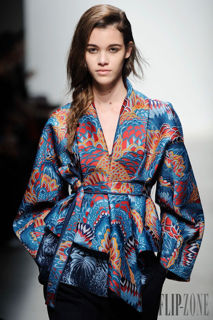 a female fashion model is showcasing a vibrant blue and red patterned kimonostyle jacket on a runway or photoshoot set. the model has a side braid hairstyle and subtle makeup.  the background is simple and white, focusing attention on the model and her attire. the jacket has a loose fit with flared sleeves and a belted waist. the overall style evokes a sense of elegance and sophistication.  the image is sharp and welllit, highlighting the intricate details of the clothing's design.