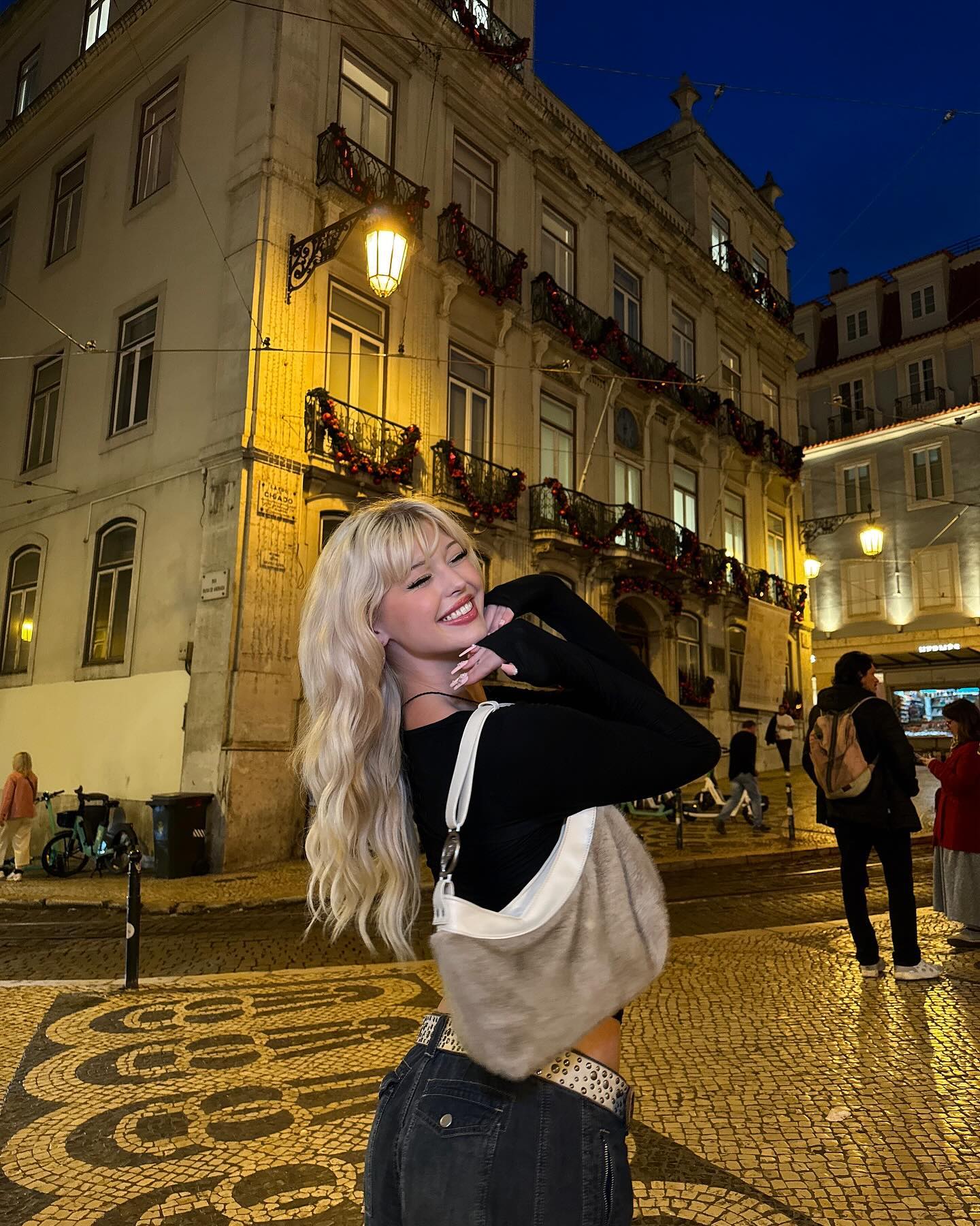 a young woman with blonde hair is posing on a cobblestone street in a city at night. she is wearing a black longsleeved top, jeans with a decorative belt, and a shoulder bag. she is smiling and has her hands near her face. in the background, there are several multistory buildings with decorated balconies and illuminated windows. a lamppost with a bright light fixture is visible on the corner of one of the buildings. other people are walking on the sidewalk in the background. the sky is a dark blue, indicating it is evening or night. the street is paved with a unique pattern of cobblestones.
