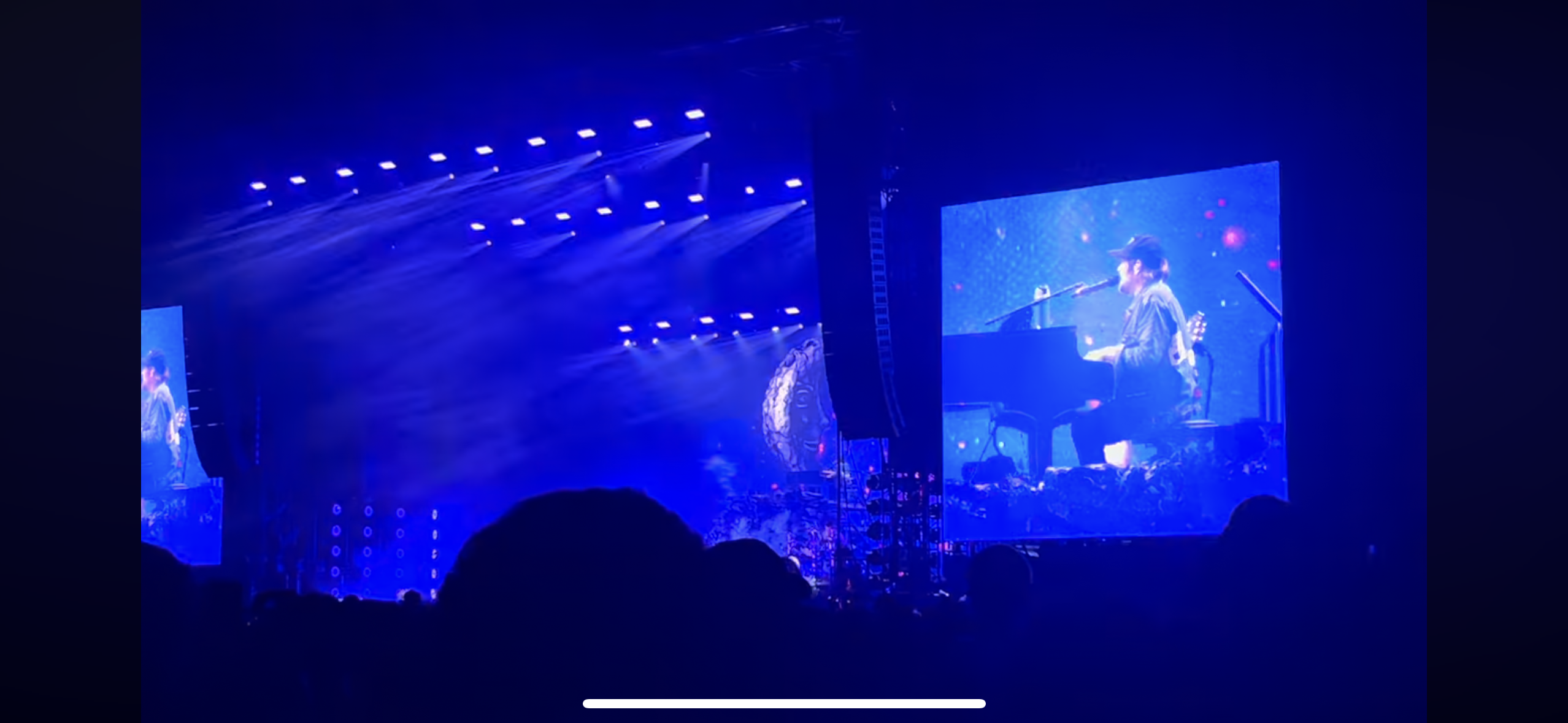 a view from the audience of a concert. the stage is bathed in blue light and a large screen displays an image of a musician playing the piano. the crowd is mostly obscured by silhouettes, but their heads are visible in the foreground.