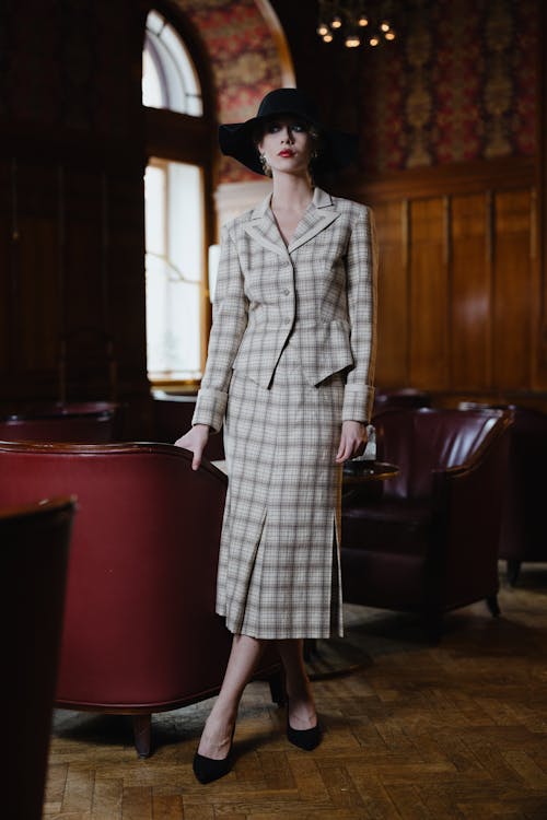 a full shot of a woman model wearing a beige and brown plaid suit with a black hat. she is standing in a room with wooden paneling and furniture, striking a pose. the outfit is elegant and retro in style.