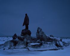 a silhouette of a person wearing a cape stands atop some rocks in a snowy landscape at night. the scene is dark and blue, with ice and snow covering the ground. a small light is visible in the sky.