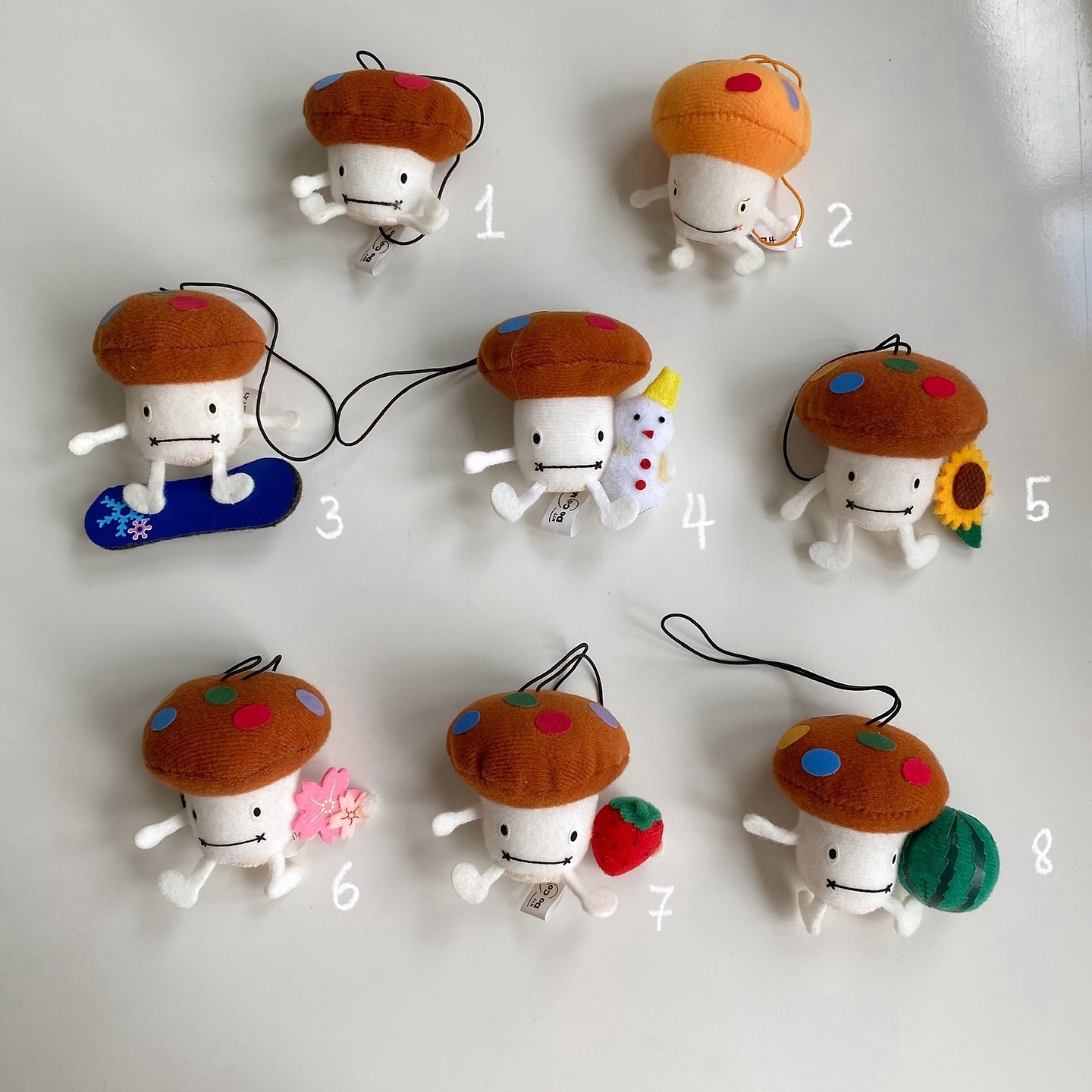 there is a collection of eight mushroom plushies on a white surface. each plushie has a white body with stitched mouth, and a cap in various colors like brown, orange, and decorated with colorful polka dots. some of them are accompanied by miniature objects: one with a snowboard, another with a snowman, a sunflower, a cherry blossom, a strawberry, and a slice of watermelon. each plushie also has a black string attached to the top of the cap. each of the mushroom plushies is labeled with a number, from 1 to 8.