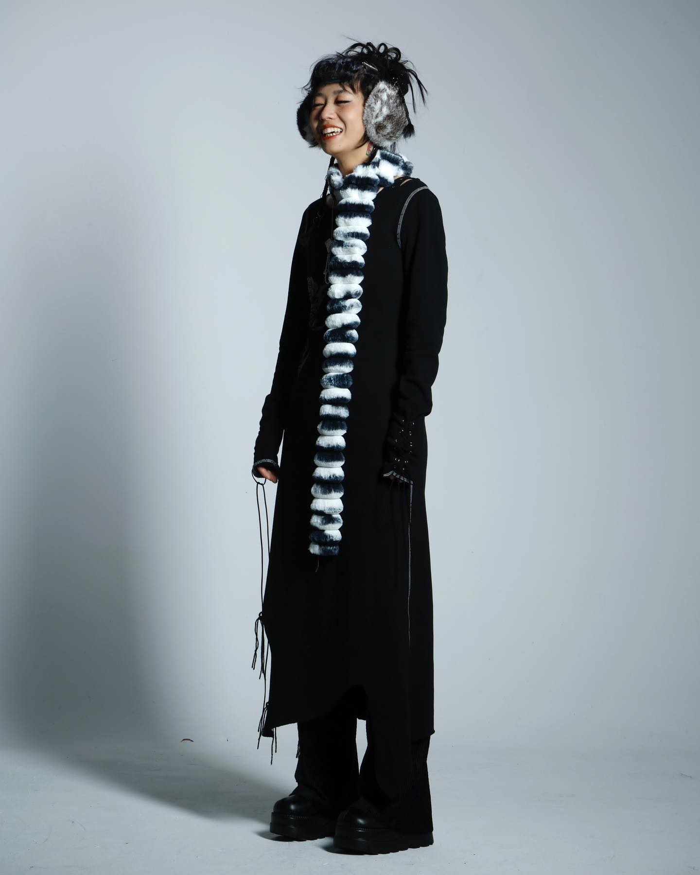 a fulllength studio shot of a young woman wearing a long black coat, a black and white striped scarf, and furry earmuffs. she is standing on a gray background and smiling at the camera. her hair is styled in a slightly messy updo. she is wearing black pants and boots. the overall style is fashionable and edgy.