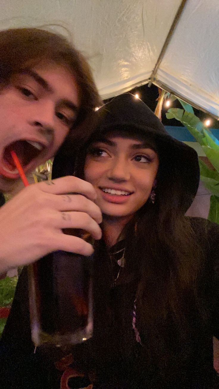 a young man and woman are smiling as they look towards the camera. the woman wears a black hoodie and has dark hair. she holds a glass in her hand and the man is holding a glass in his hand. they are indoors and it is dark, with a red straw visible in the man's glass.