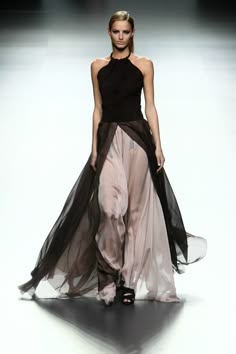 a beautiful woman walks down a runway in a long, flowing dress. the dress is black and white, with a sheer top layer that shows off her figure. she has long, dark hair and is wearing minimal makeup. the image is a classic example of high fashion, and the woman is a striking example of elegance and beauty.