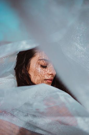 a close up portrait of a woman with a soft, dreamy expression. her face is partially hidden by a delicate lace fabric, creating a sense of mystery and intrigue. the fabric is soft and flowing, and the woman's hair is long and dark, framing her face. the image is soft and ethereal, with a romantic and timeless quality.  the woman's skin is flawless, and her eyes are closed, as if she is lost in thought. the lighting is soft and diffused, creating a dreamy and romantic atmosphere. the image is evocative and suggestive, inviting the viewer to create their own story about the woman and her thoughts.