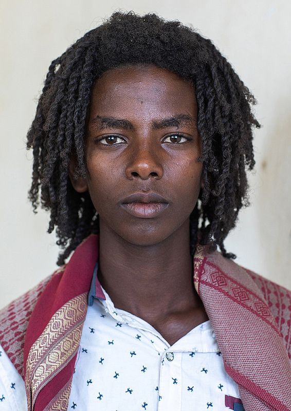 a closeup portrait of a young man with dark skin and dreadlocks. he is wearing a white shirt with a dark pattern and a red shawl with gold detailing. he has a serious expression on his face.