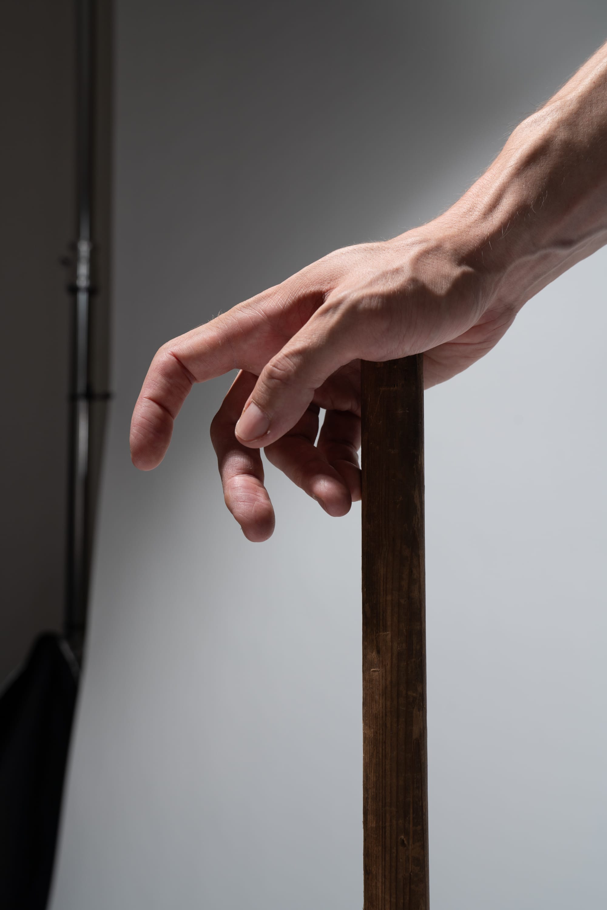 a closeup shot of a man's hand gripping a wooden stick. the man's arm is extended, and the stick is held vertically. the background is a plain white wall, and the lighting is soft and diffused, typical of a studio setting.