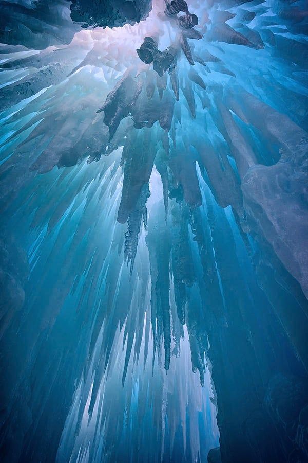 a view from inside an ice cave with long, sharp icicles hanging from the ceiling, illuminated by a blue and white light.