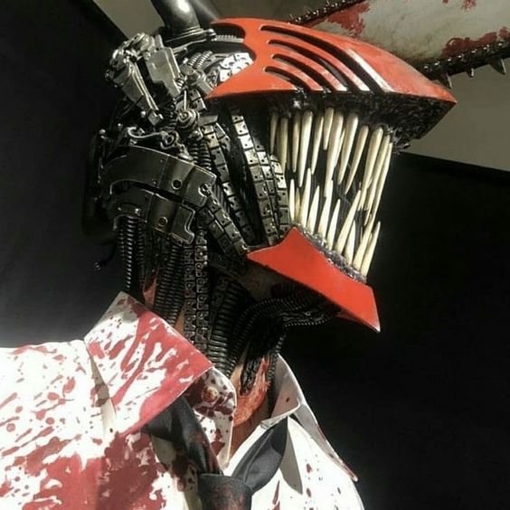 this image shows a person in a chainsaw man cosplay. they are wearing a red helmet with large teeth and a chainsaw blade on top, with intricate mechanical details visible. the helmet covers the top half of their face. they are also wearing a white shirt heavily splattered with fake blood. the background is dark.