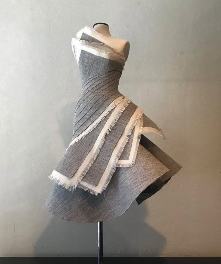 the image shows a stylish grey dress with white trim on a mannequin, set against a pale wall in a studio setting. the dress features an asymmetrical design with a fitted bodice and a layered, flared skirt, emphasizing its unique textile design and fashionforward aesthetic.