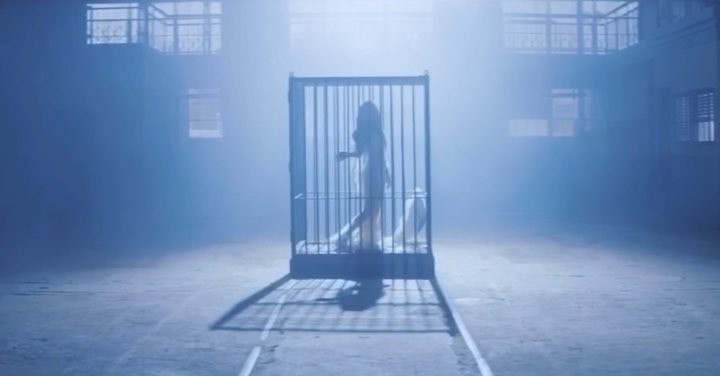 a woman is inside a cage in a large, empty, bluetoned space. the cage casts a long shadow on the ground. there are buildings with windows visible in the background, suggesting an industrial or institutional setting. the overall atmosphere is surreal and conveys a sense of confinement.