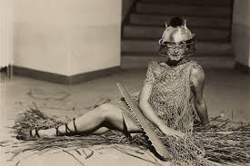 a black and white photograph of a woman in a surreal pose. she is wearing a costume that incorporates a mesh or net fabric and a helmetlike mask that partially obscures her face. the woman is sitting on a floor covered with straw or grass.  a large saw is positioned beside her, adding to the unusual and mysterious feel of the image. the overall style is artistic and evocative.