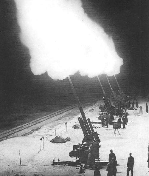 a black and white photograph depicting a battery of german 8.8 cm flak guns firing at night during world war ii.  the bright flashes and smoke from the explosions illuminate the scene, showing the guns and soldiers manning them. the setting appears to be a snowy or icy field near a railway line.  the image is a historical record of antiaircraft defenses in action.
