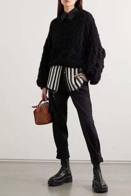 a full shot shows a woman standing in a studio wearing a black cable knit sweater layered over a black buttondown shirt and a black and white striped shirt. she is also wearing black jeans cuffed at the ankles and black platform boots. she is holding a small brown purse in her left hand. the background is white.