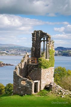 an eyelevel view of a weathered stone ruin of a tower perched on a hill overlooking a body of water. the tower features arched windows and a staircase leading to an upper level. lush green ivy climbs the stone walls. the background showcases a picturesque scene with blue water, a distant city, and rolling hills under a cloudy sky.
