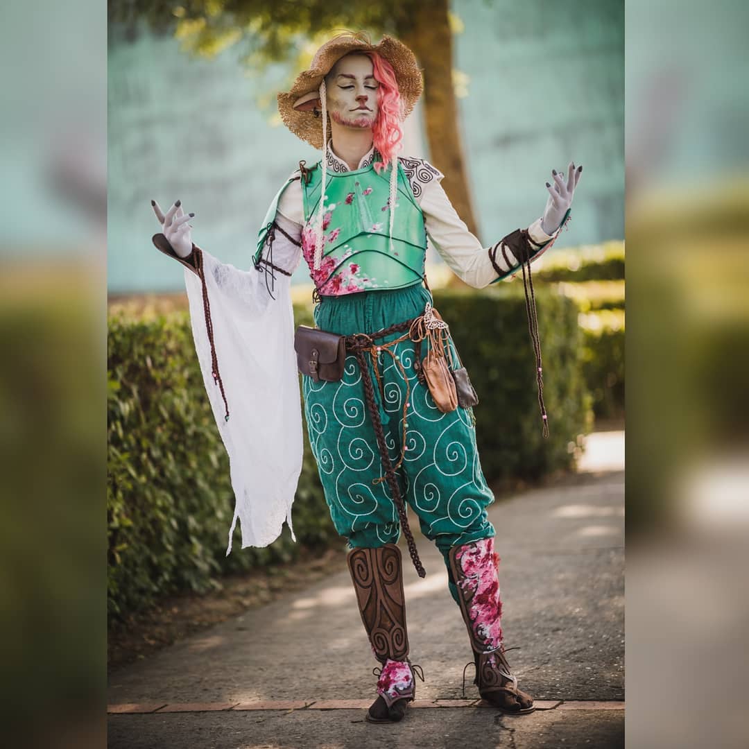 the image shows a woman in an elaborate cosplay costume. she's dressed as a fantasy character, possibly an elf or halfling, reminiscent of a character from a game like dungeons & dragons or a similar setting. her outfit features a green and pink floralpatterned breastplate, teal pants with swirling designs, and brown leather boots with pink floral accents. she wears a straw hat and has pink hair. the overall style suggests a ranger, druid, or similar naturebased class.  she's outdoors, on a path in what seems to be a park, standing with a relaxed and peaceful pose, hands outstretched slightly.  the costume is highly detailed and wellcrafted.