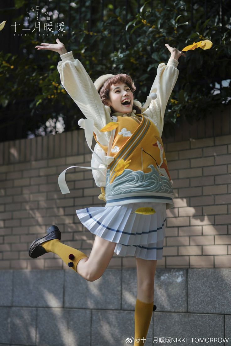 a young asian girl wearing a white and yellow sweater with a pleated white skirt and black shoes jumps in the air while smiling. she has on yellow knee high socks. the background is a brick wall with leaves and greenery visible. she is also holding yellow leaves in her hands.
