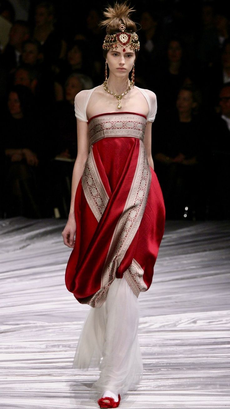 a model walks the runway wearing a stunning red and white dress inspired by indian sari designs. the dress is a flowing red silk with a white underlayer and ornate silver detailing. the model is adorned with elaborate gold and jeweled headpiece and necklaces.  the background is blurred, focusing attention on the model and her attire. the overall scene suggests a high fashion runway show.