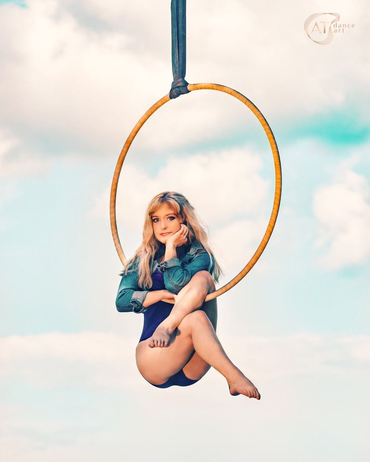 a young woman sits relaxed in a blue denim jacket and a blue leotard in an aerial hoop. she is posed in a seated position with her legs and arms bent, her chin on her hand, staring directly at the camera. she is outdoors with blue skies and white clouds behind her. the bright sunlight illuminates the scene and makes the colors pop.