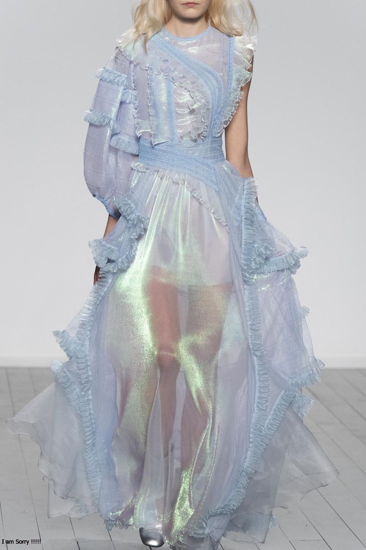 a model wearing a light blue, sheer, ruffled dress walks down a runway. the dress is iridescent and has a delicate, feminine feel. the model is wearing her hair down and has a natural makeup look. the background is a white wall, which allows the dress to be the focal point of the image.
