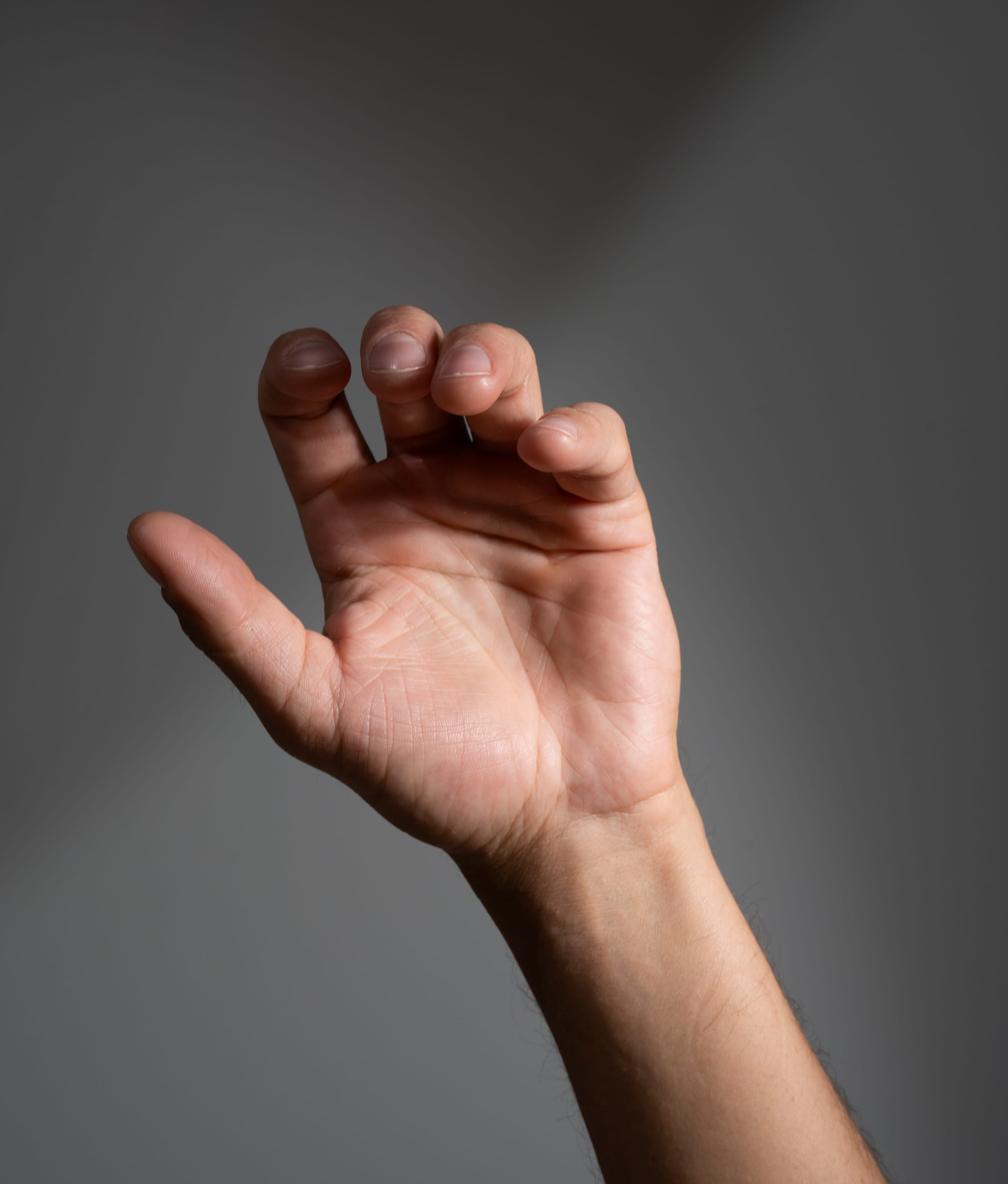 a man's right arm and hand are reaching up against a grey background. the hand is partially open, as if reaching for something above. strong artificial light creates shadows on the hand and background.