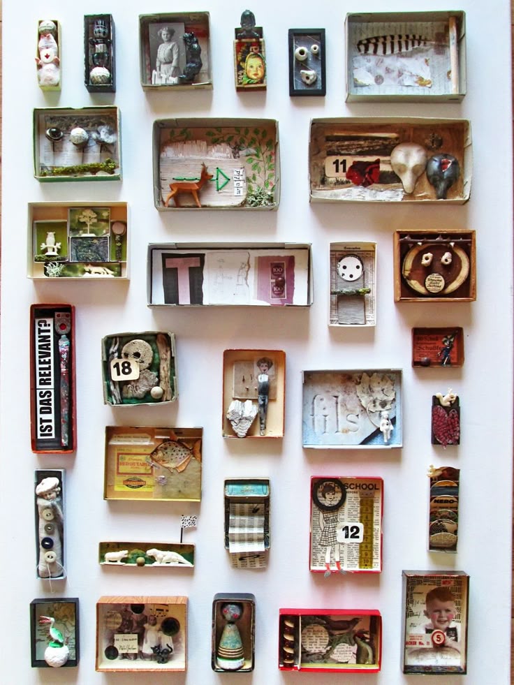 the image shows an arrangement of small shadow boxes, each containing a unique collage or assemblage of found objects and mixed media elements. the boxes are diverse in size, shape, and color, and they are displayed on a white background, presumably a wall. the contents of each box vary, including items such as figurines, paper cutouts, numbers, letters, small sculptures, and other miscellaneous objects. the overall effect is a collection of miniature artworks, each telling its own story or evoking a specific mood.