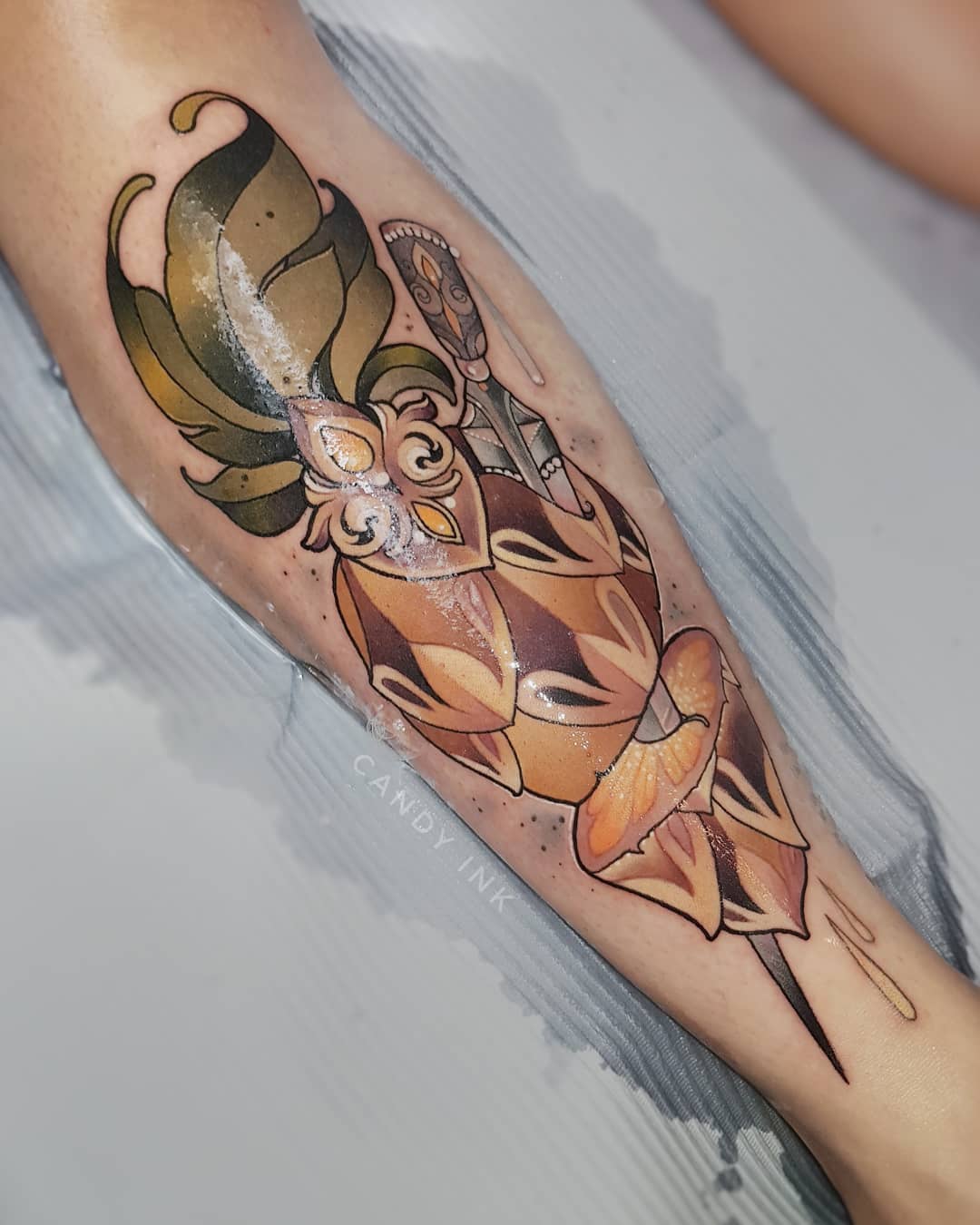 a person's leg with a freshly done neotraditional style tattoo of a stylized flower with a dagger through it.  the tattoo has brown, green, and yellow tones and is outlined in black. the tattoo is still covered in a layer of ointment.