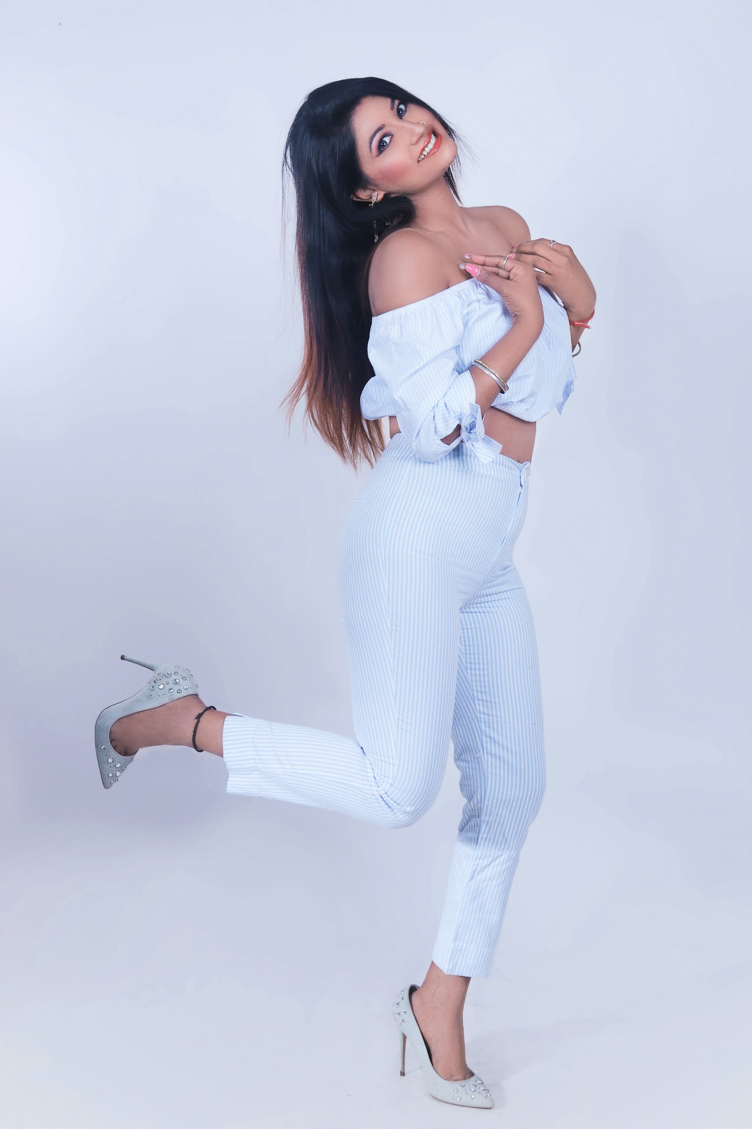a young, beautiful woman is showcasing a stylish outfit in a studio shot against a white background. she's wearing a light blue offtheshoulder top and matching striped pants.  her pose is playful and confident, one leg lifted slightly, and she is smiling at the camera. the overall aesthetic is modern, chic and fashionable. her accessories include highheeled shoes and jewelry.  the image is welllit, highlighting the details of the outfit and the model's features.