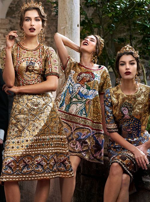 hree models are posing in ornate, gold dolce & gabbana dresses.  the dresses feature intricate embroidery and embellishments, including sequins and beads, and depict religious byzantineinspired imagery. each model wears a jeweled crown or tiara and coordinating jewelry. the setting is outdoors, with a stone column in the background. the overall aesthetic is one of luxury, elegance, and regal sophistication.