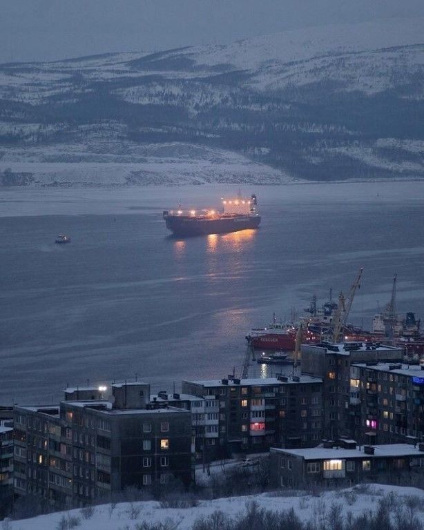 the image depicts a twilight scene in a northern russian port city, possibly murmansk, situated on the barents sea.  the foreground shows a cluster of multistory apartment buildings covered in snow. in the middle ground, a harbor is visible with several vessels, including cargo ships and cranes.  a large tanker ship with illuminated lights is positioned in the waterway further out. the background features snowcovered hills or low mountains stretching across the horizon. the overall ambiance is cold, dark, and peaceful, indicative of a harsh winter climate. the lighting suggests it is either early morning or late evening.