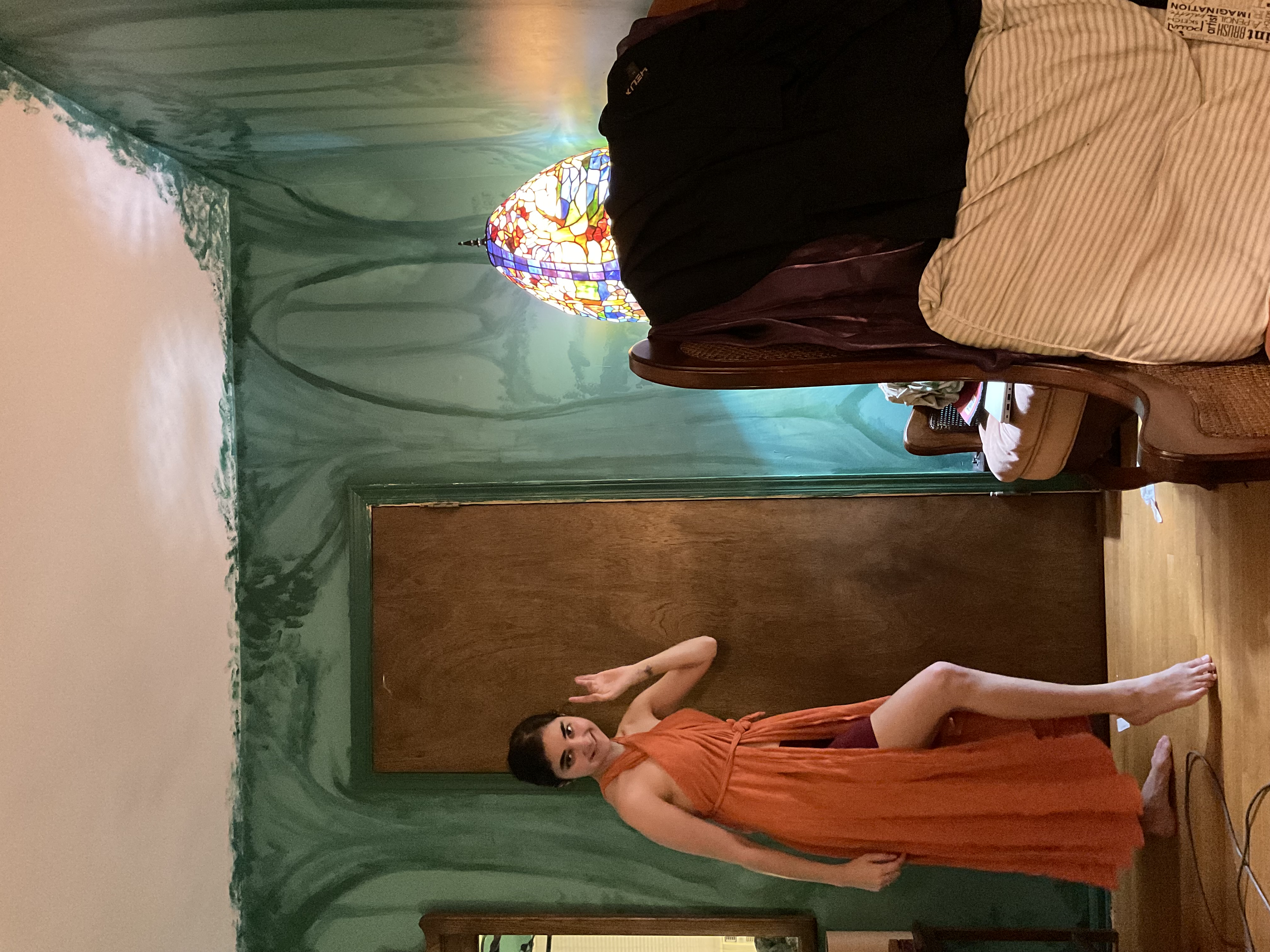 a young woman in a flowing orange halter dress with a high slit stands in a room with uniquely painted green walls depicting a forest scene. she is posing, waving gently. the room also features a wooden door, an upholstered armchair with wicker details, and a tiffanystyle stainedglass lamp. the overall aesthetic is eclectic and bohemian.