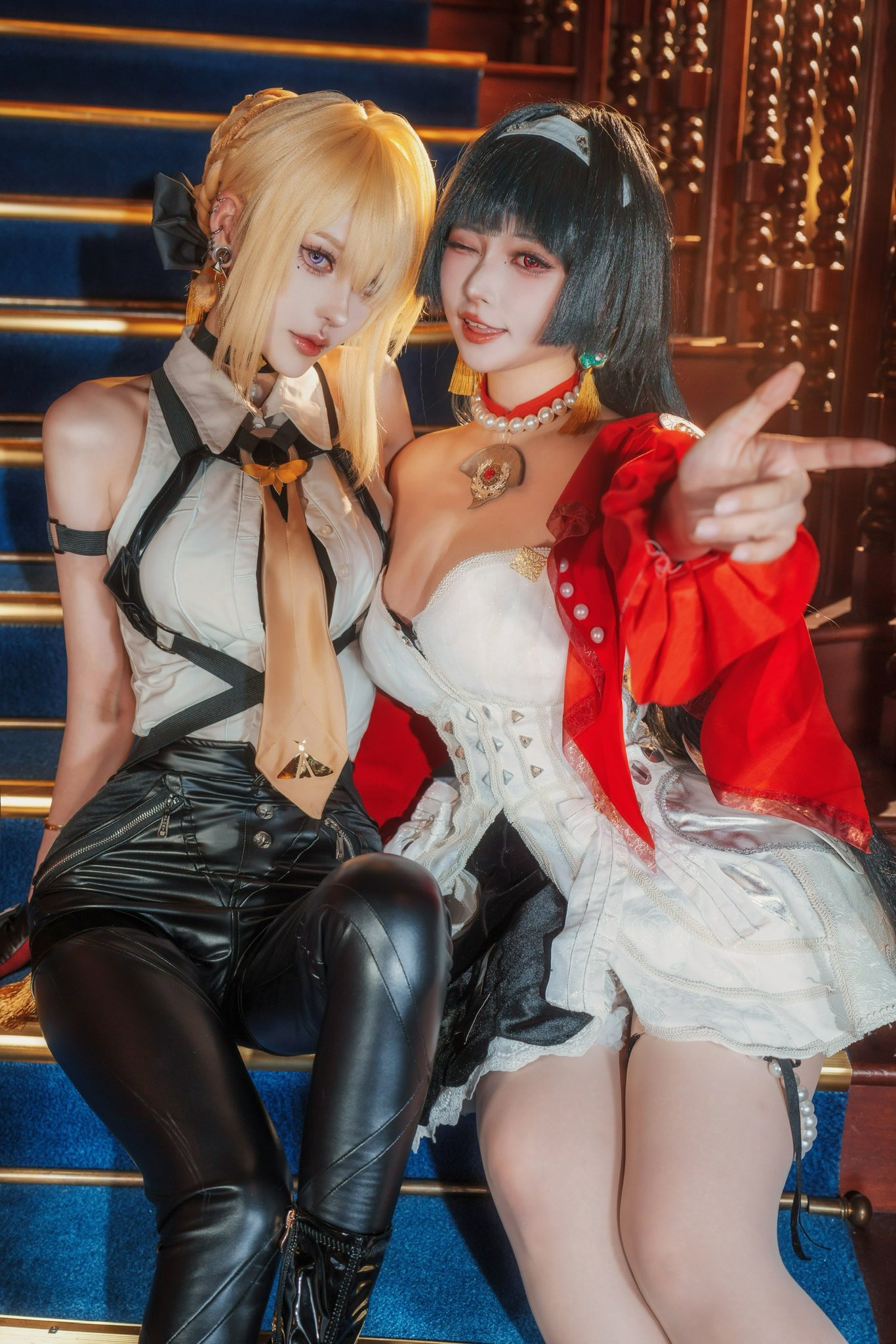 two women in elaborate cosplay costumes are sitting on a staircase. one is dressed as a blondehaired character in a leather outfit with a white shirt and tie. the other is dressed in a white and red dress with a corset. they are smiling and posing for the camera. the setting appears to be indoors in a grand building.