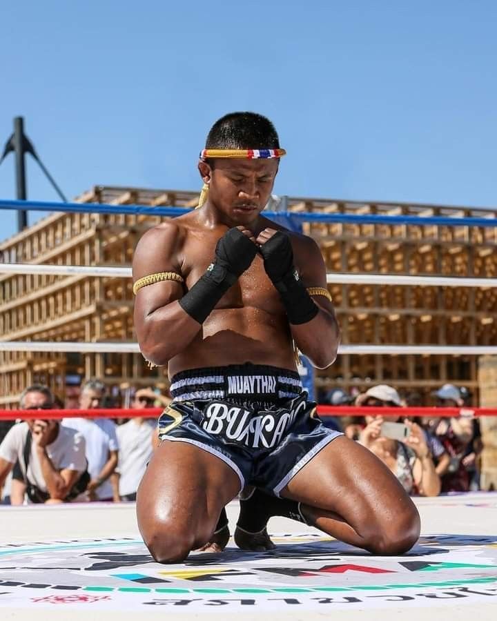 a muay thai fighter kneels in the ring, his head bowed in prayer, hands clasped together in front of his chest. the image captures the intensity and focus of the fighter as he prepares for battle, showcasing the discipline and dedication required for martial arts. his physique is sculpted and strong, reflecting years of rigorous training. the fighter's traditional muay thai shorts and headband add to the authenticity of the scene. the image is a powerful representation of the spirit of a warrior, ready to face any challenge.