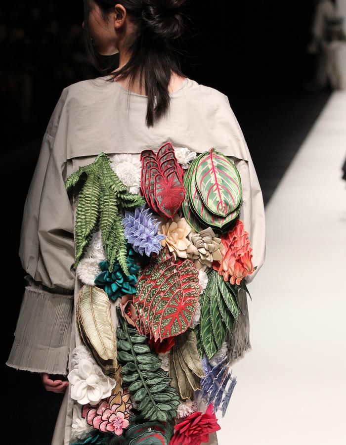 a model walks down a runway in a unique and creative outfit. the outfit is primarily a beige shirt with a large floral and leaf design embroidered onto the back. the floral and leaf design is very detailed, intricate, and realistic. the model's long dark hair is pulled back, and they are not looking at the camera. the background is white and blurred.