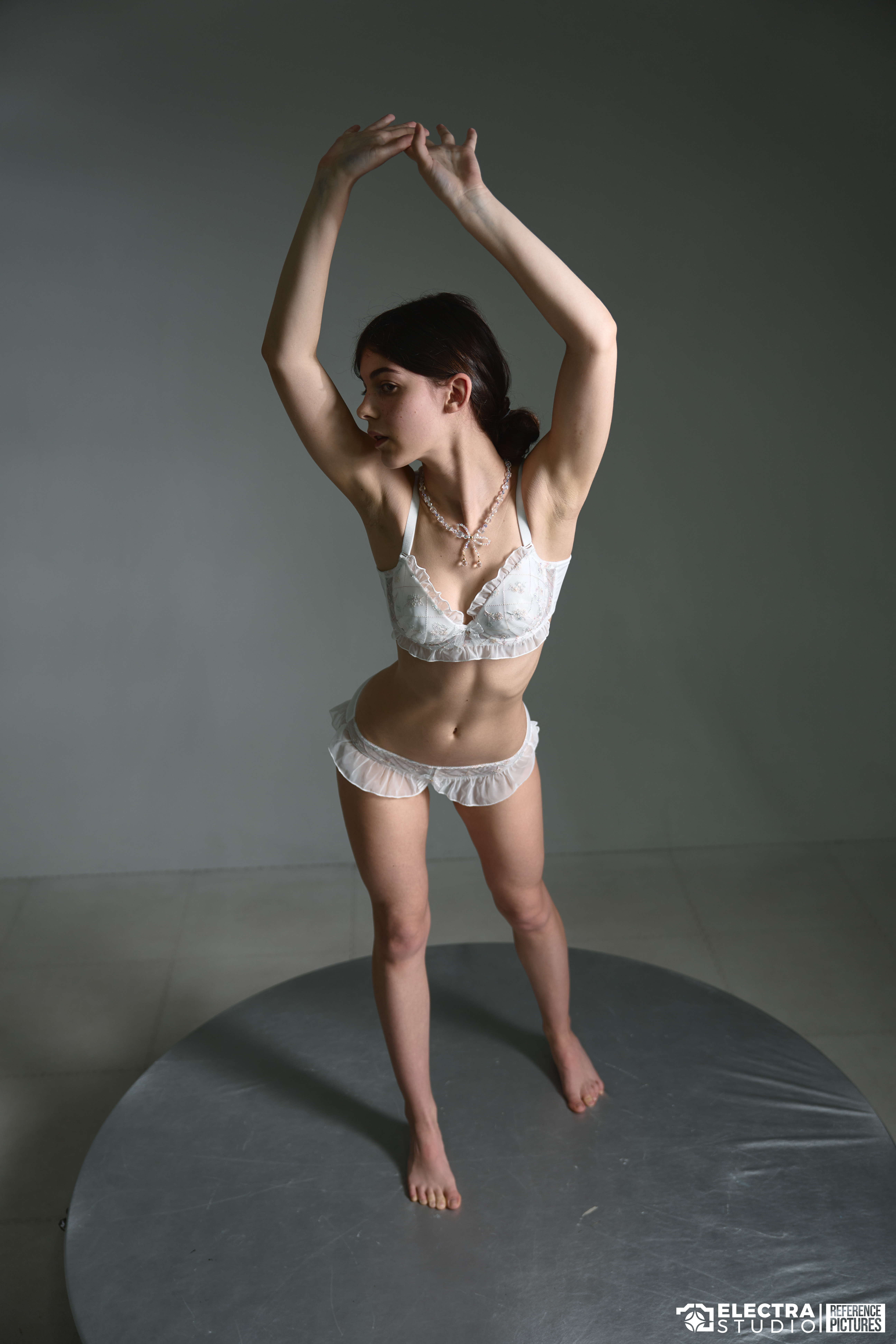 a fullbody studio shot shows a young woman wearing white lingerie and a necklace, standing barefoot on a grey circular platform, with her arms raised above her head against a light grey background.