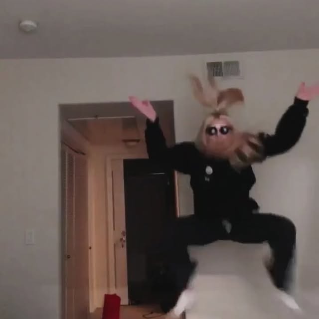 a woman is jumping in the air indoors. she is wearing a black outfit and sunglasses. her blonde hair is flying upwards. there is a doorway behind her and it looks like she is in a home or apartment.